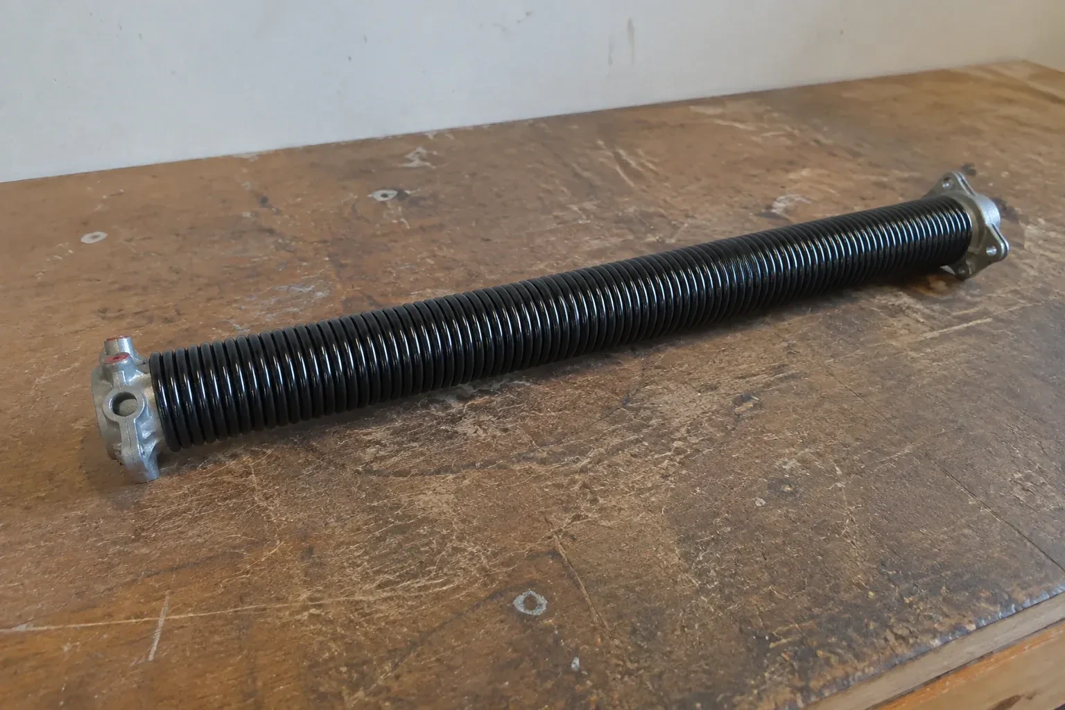 Detached black coil spring with metal end fittings on a workbench. — Torsion Spring Replacement, Niagara Falls