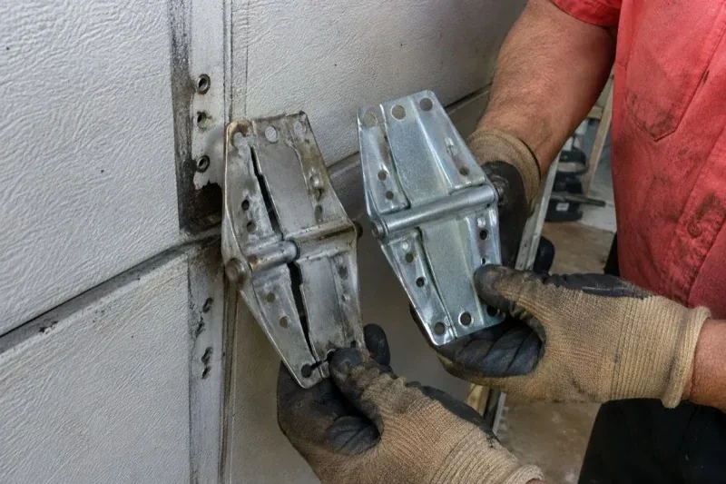 Gloved hands holding two metal garage-door hinges side by side, one discolored with a visible crack and the other bright silver. — Garage Door Hinge Replacement, Niagara Falls