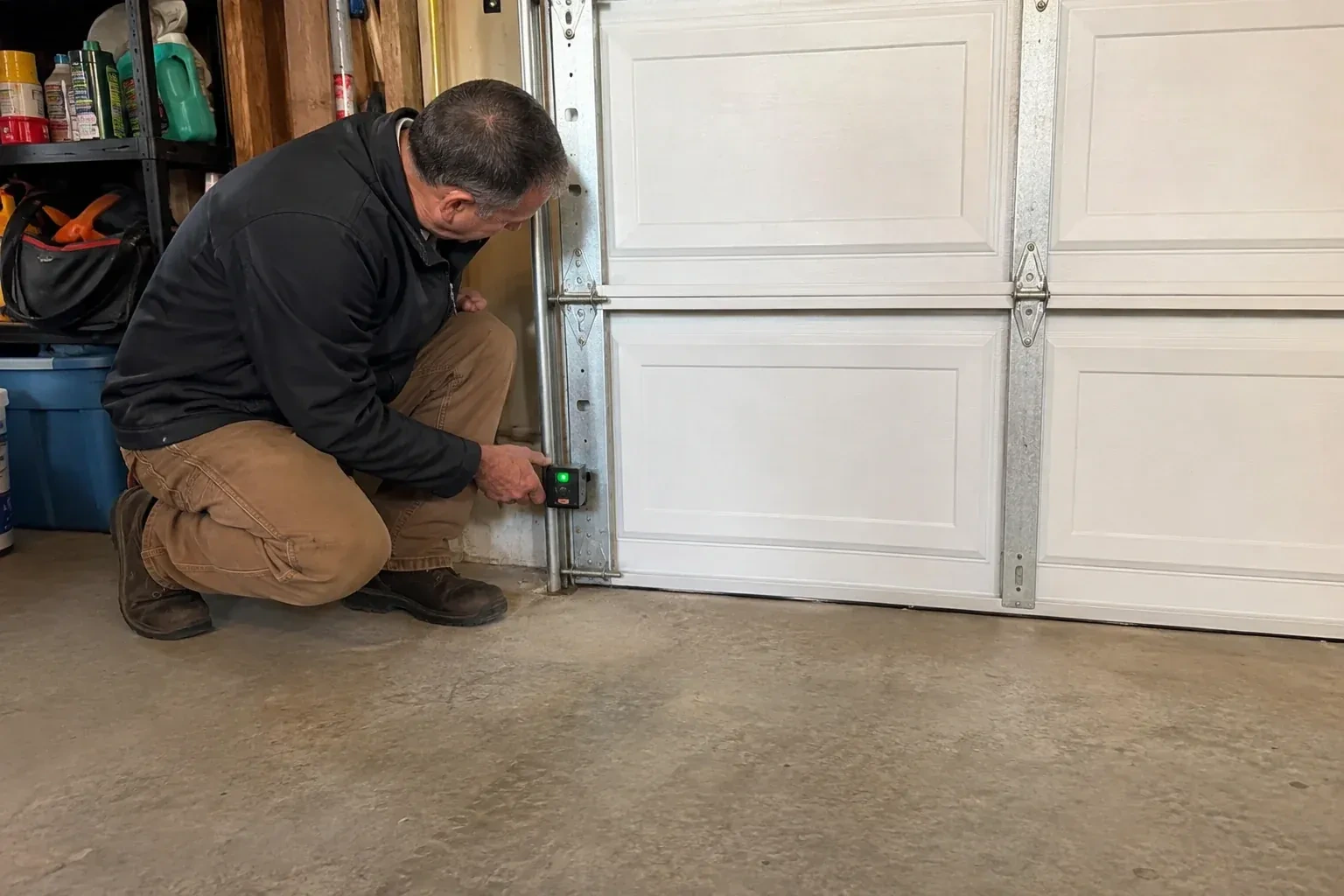 Technician kneeling beside a white paneled garage door, holding a small black box with a green light against the vertical track. — Photo-Eye Sensor Alignment or Replacement, Niagara Falls