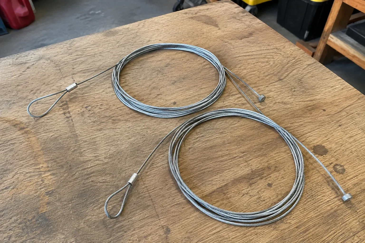 Two coiled steel cables with loop and stop ends on a workbench. — Garage Door Cable Replacement, Chippawa